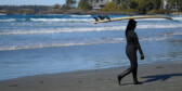 A female surfer in a wet suit walks on the shore carrying a surfboard over her head.