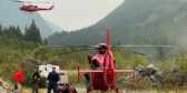 One red helicopter sits on the ground while people get ready to fly it. Another red helicopter has just taken off.