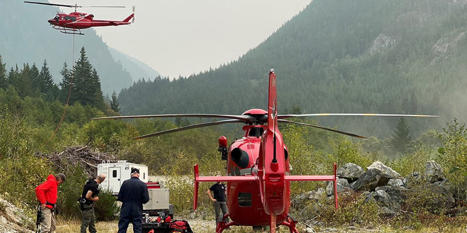 One red helicopter sits on the ground while people get ready to fly it. Another red helicopter has just taken off.