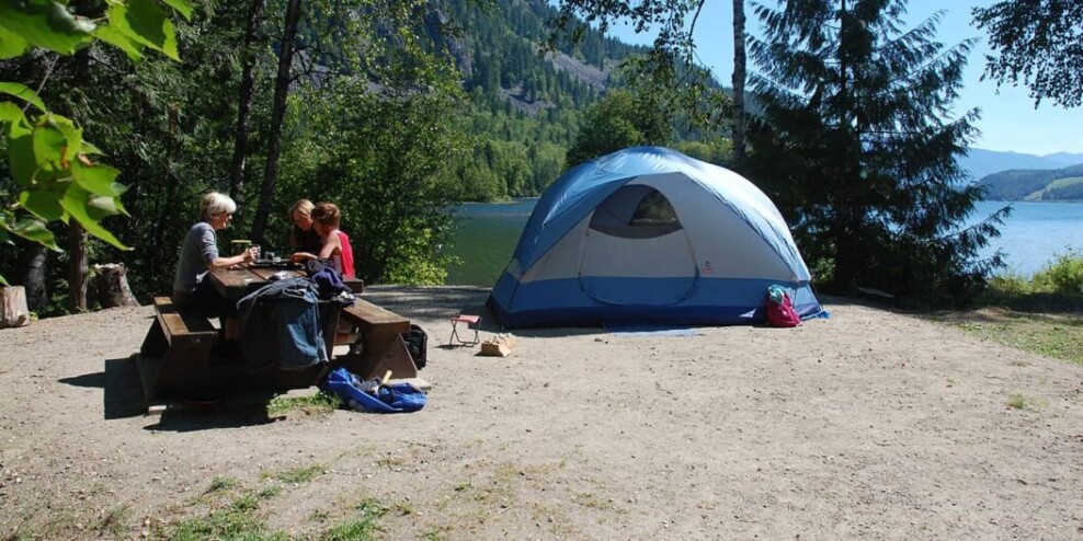 Three people sit at a picnic table next to their dome tent. There are trees and mountains in the background. It's a sunny day.