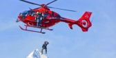 A red helicopter lowers someone onto a mountain on a sunny day.