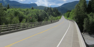 A picture of Highway 19 going off into the distance on a sunny day. One person is driving, another is walking on the sidewalk.