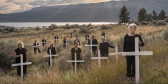 A group of mothers stands in a field. Each mother stands behind a white cross to commemorate a child lost to drugs.