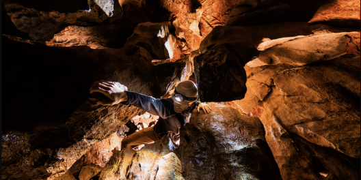 A man in a head lamp and helmet climbs through a small crevice in a dark karst cave.