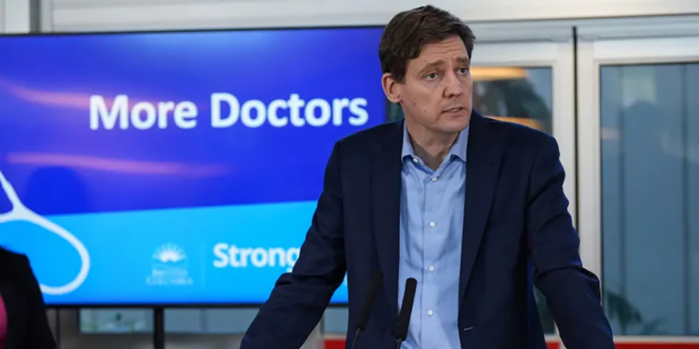 David Eby stands in front of a sign that reads "More Doctors."